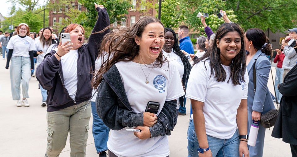 Students in white CWRU alum shirts with big smiles as they walk during clap out with members of the community cheering in the background
