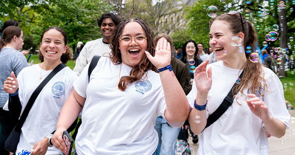 Three students dressed in white CWRU alum shirts smile while walking during clap out while bubbles waft past