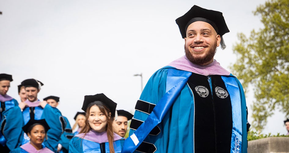 A graduate from the School of Dental Medicine smiles as he walks across campus with others in a line behind him