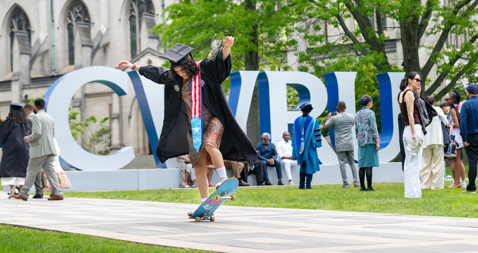 A graduate in their cap and gown rides a skateboard down the binary walkway with the CWRU letters in the background.