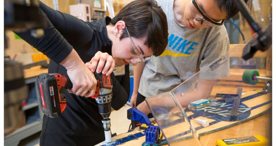 Two individuals in safety goggles work intently on a project. One drills a hole into a blue skateboard clamped to a table, focusing on precision.