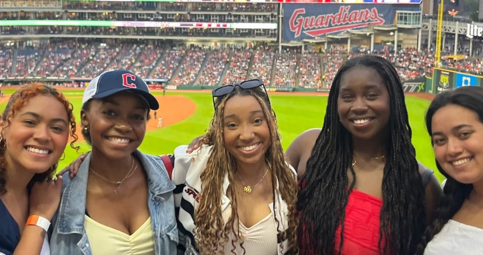 Five women smile at the camera inside a baseball stadium with the diamond behind them.