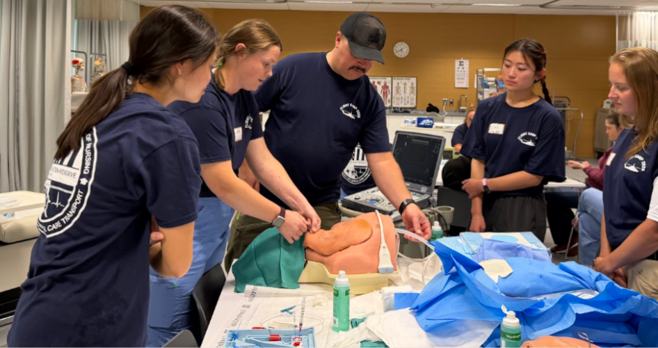 Students in dark blue shirts stand around a medical mannequin.