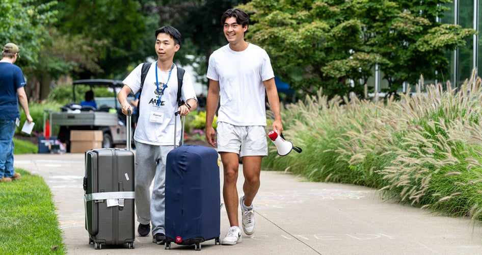Two students walk down a path as with suitcases during move-in