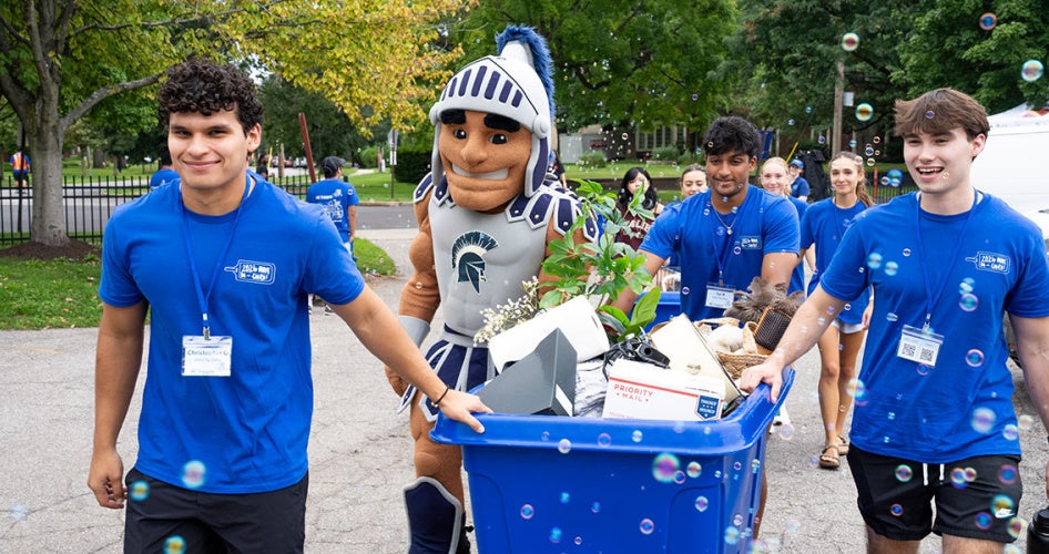 Student orientation leaders move a cart full of a new student's belongings in using a cart as bubbles blow on them and Spartie walks alongside