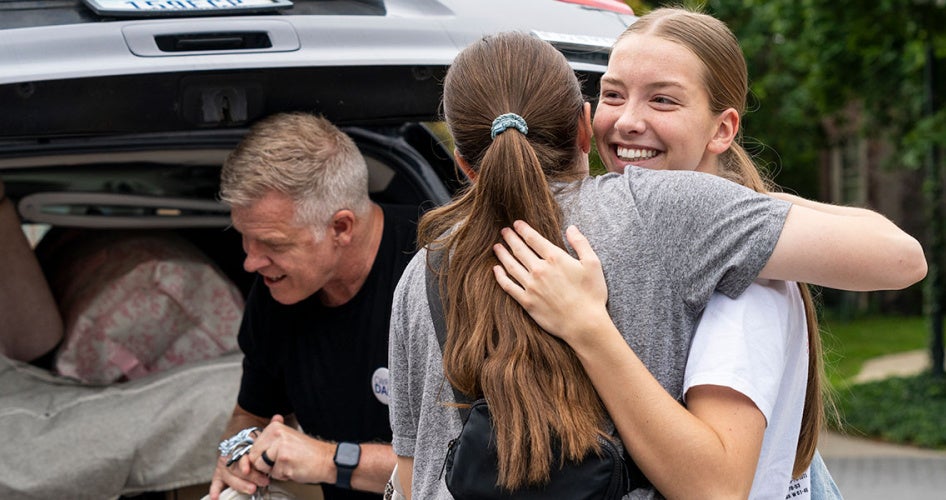 An arriving student hugs a family member as another works on unloading items from their trunk