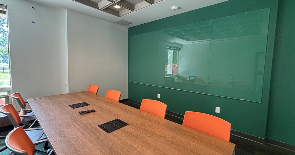 Photo of an empty meeting space with a table surrounded by orange chairs and a teal wall with glass panel for writing on