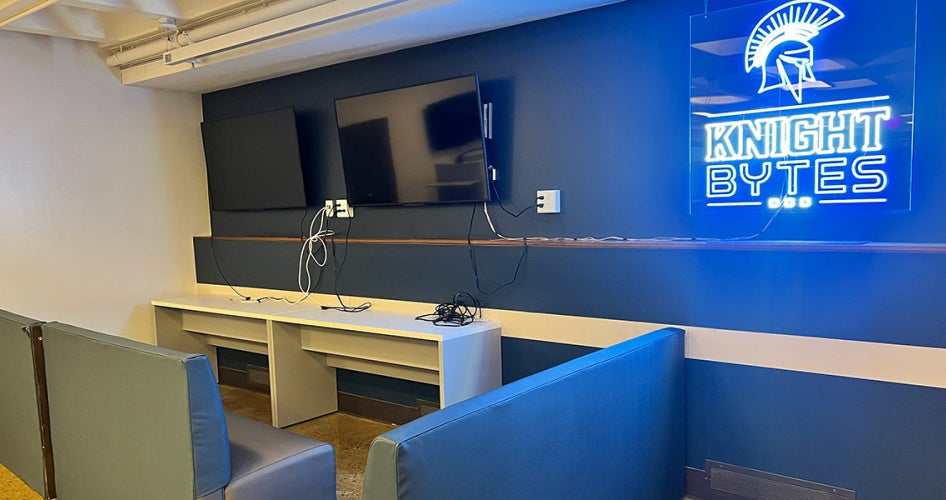 A neon Knight Bytes sign is illuminated next to a TV with couches nearby