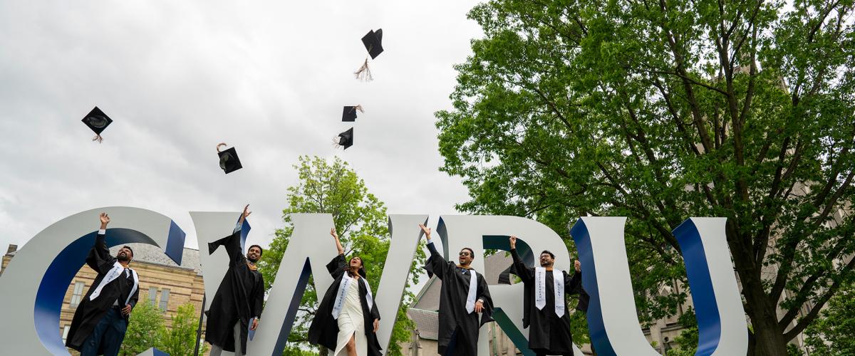 Five students stand in front of the CWRU sign and throw their commencement hats in the air