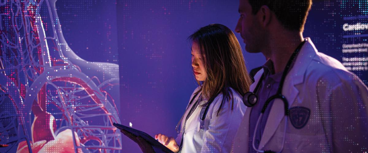 Two Case Western Reserve University medical students stand in a dark room looking at a hologram of a body on a wall
