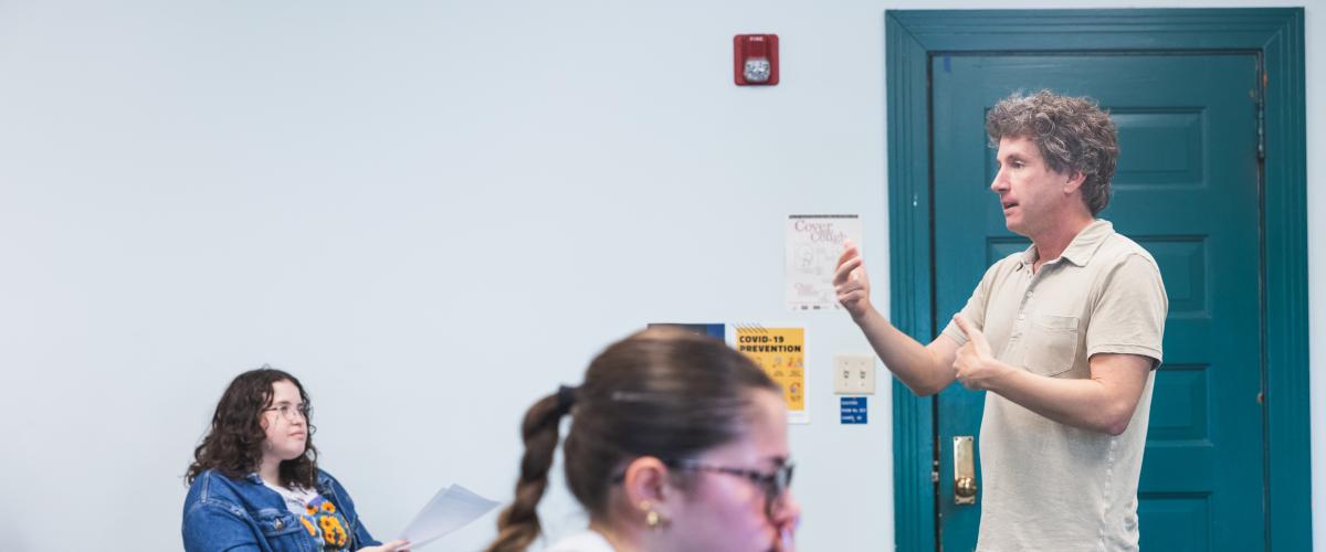 A classroom setting with a teacher standing and speaking. Two students, one focused on papers and the other on a laptop, listen attentively.