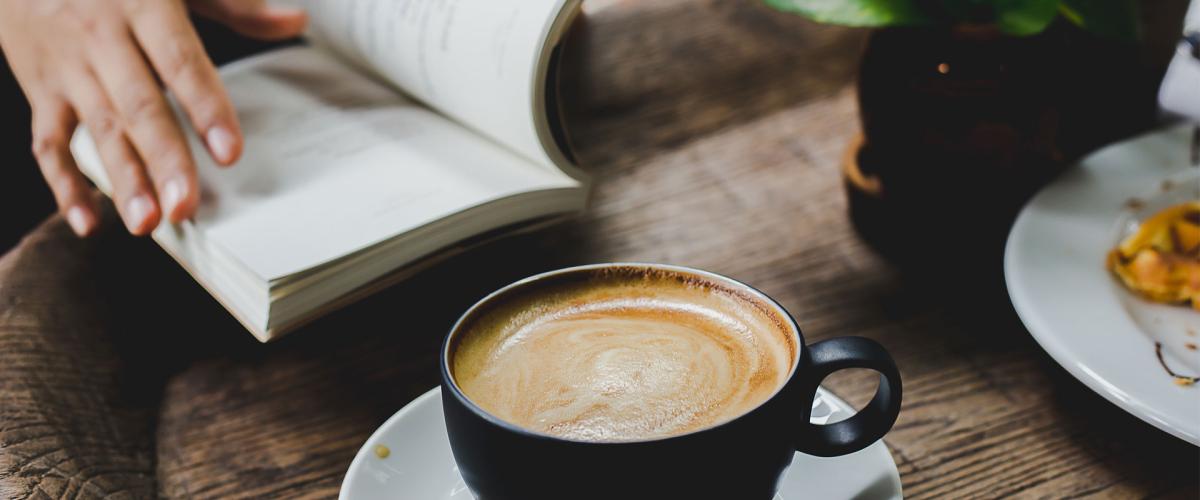 Close-up photo of coffee in a cup on a table and someone reading a book next to it 