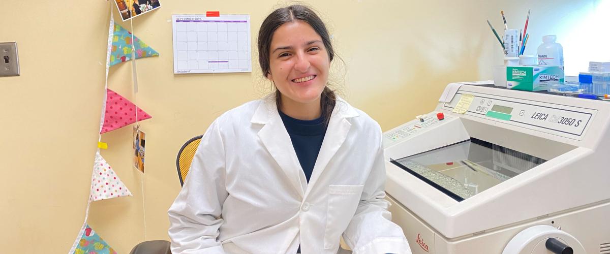 A woman wearing a lab coat and blue gloves smiling in a lab. She is next to scientific equipment.