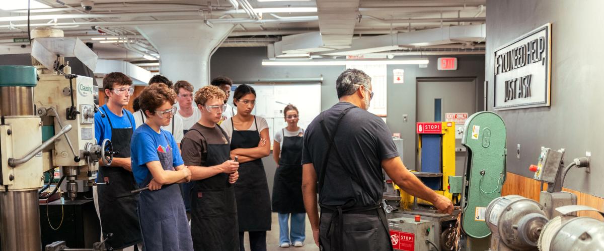 A group of students wearing safety goggles and black aprons attentively observes an instructor operating a machine.