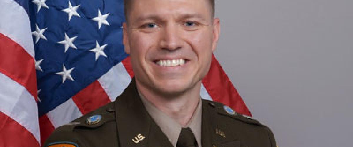 Person wearing military uniform, smiling in front of American flag