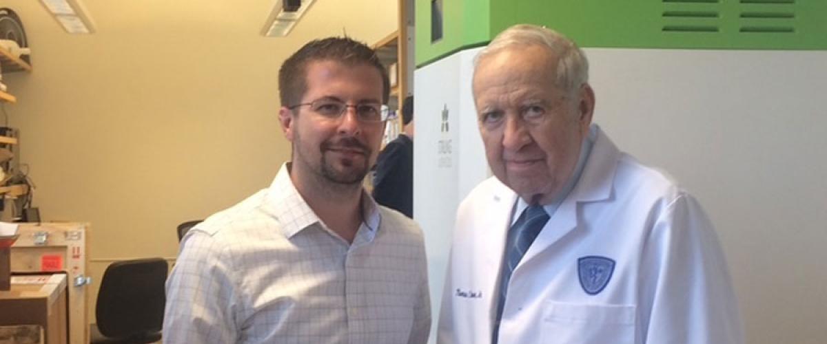 Two men standing side by side, one on left wearing dress shirt and on right white lab coat