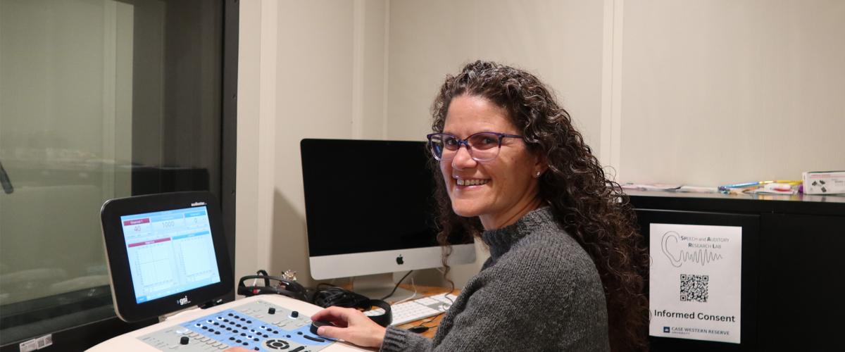 A woman sits with hearing assessment equipment in a sound-isolated booth