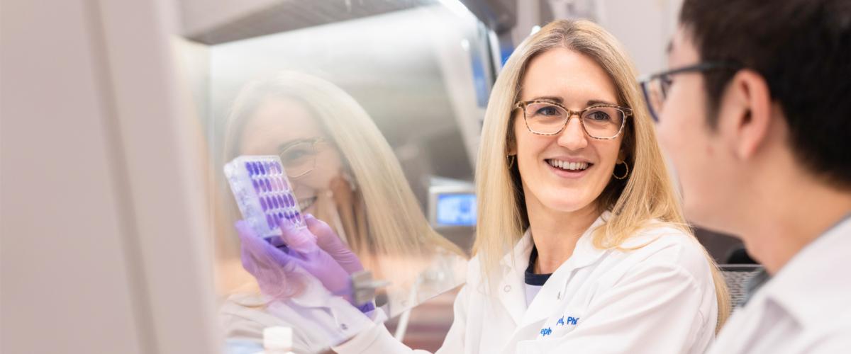 A woman in a lab coat and gloves smiles while holding a test tube rack, engaging with a colleague.