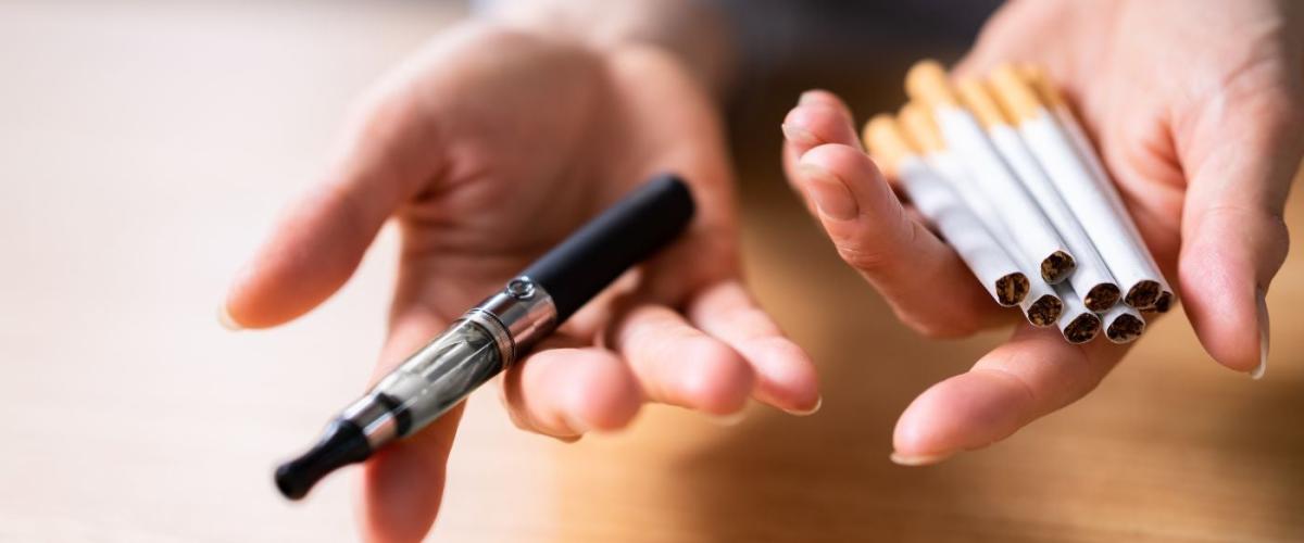 Hand Holding Vape And Tobacco Cigarettes Over Wooden Desk