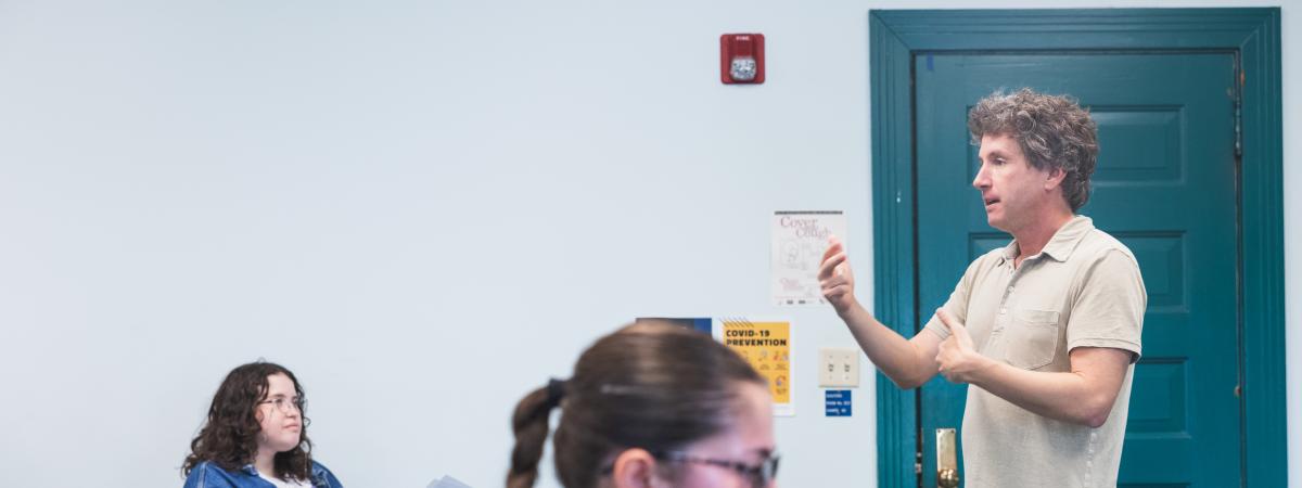 A classroom setting with a teacher standing and speaking. Two students, one focused on papers and the other on a laptop, listen attentively.