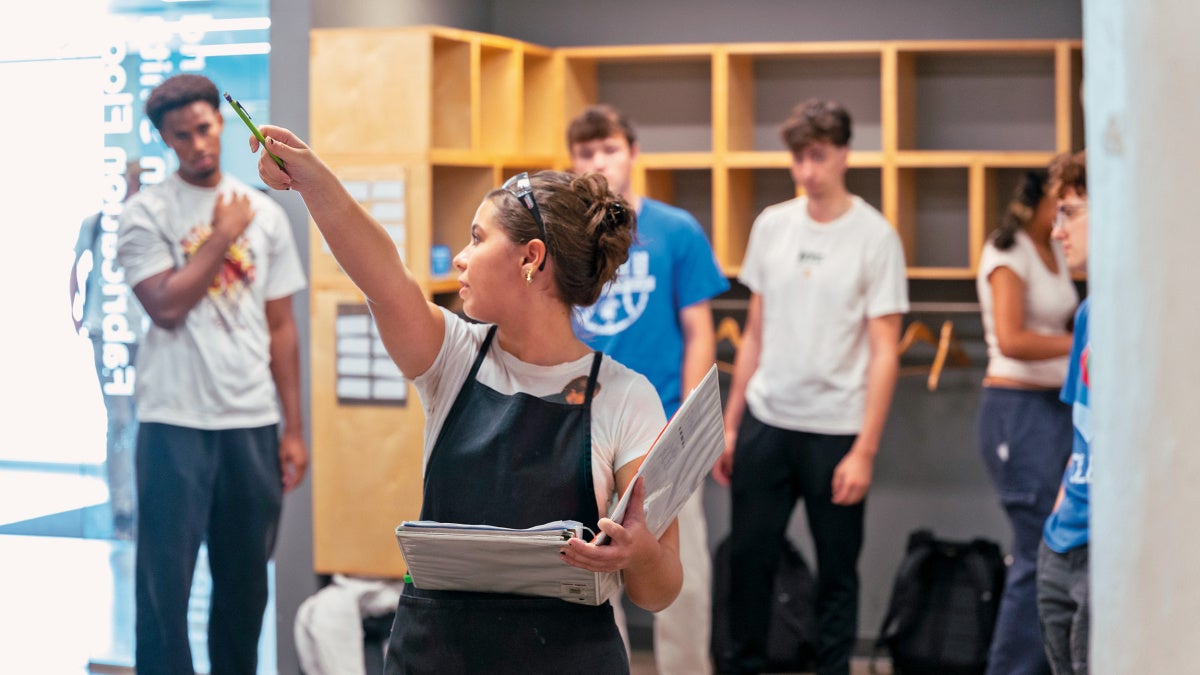 A student  in a black apron points with a pen, holding a notebook, in a room with wall cubbies. Several other students watch attentively.