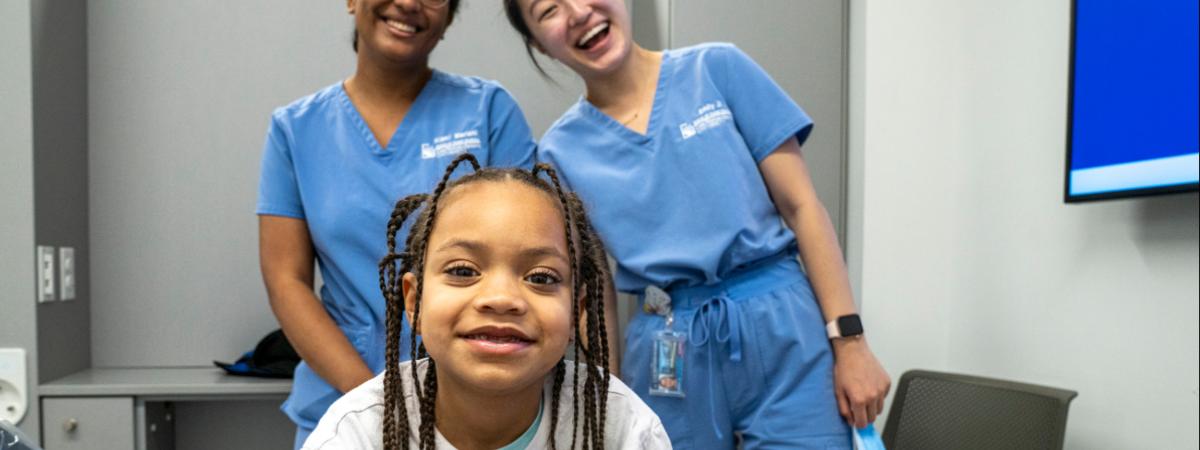 A young girl with braids in a white shirt poses in front of two dental students.
