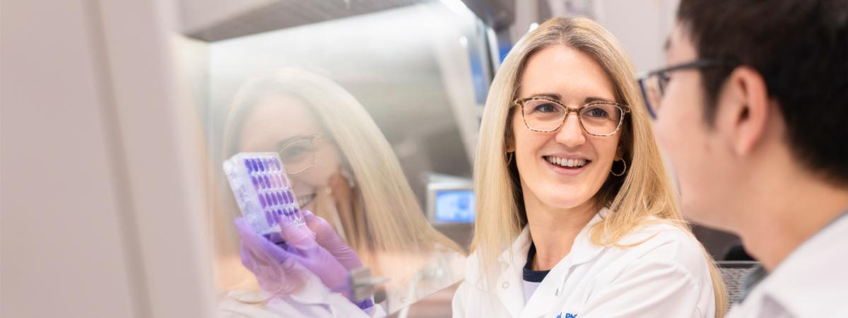 A woman in a lab coat and gloves smiles while holding a test tube rack, engaging with a colleague.