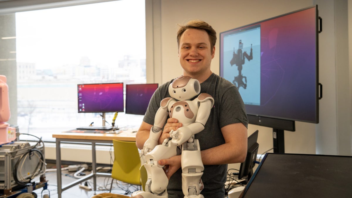 Austin holds a robot in front of a large wall-mounted screen displaying a robot silhouette
