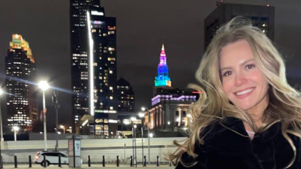 Caroline Deyoe in black coat stands smiling in front of the Cleveland skyline lit up at night.