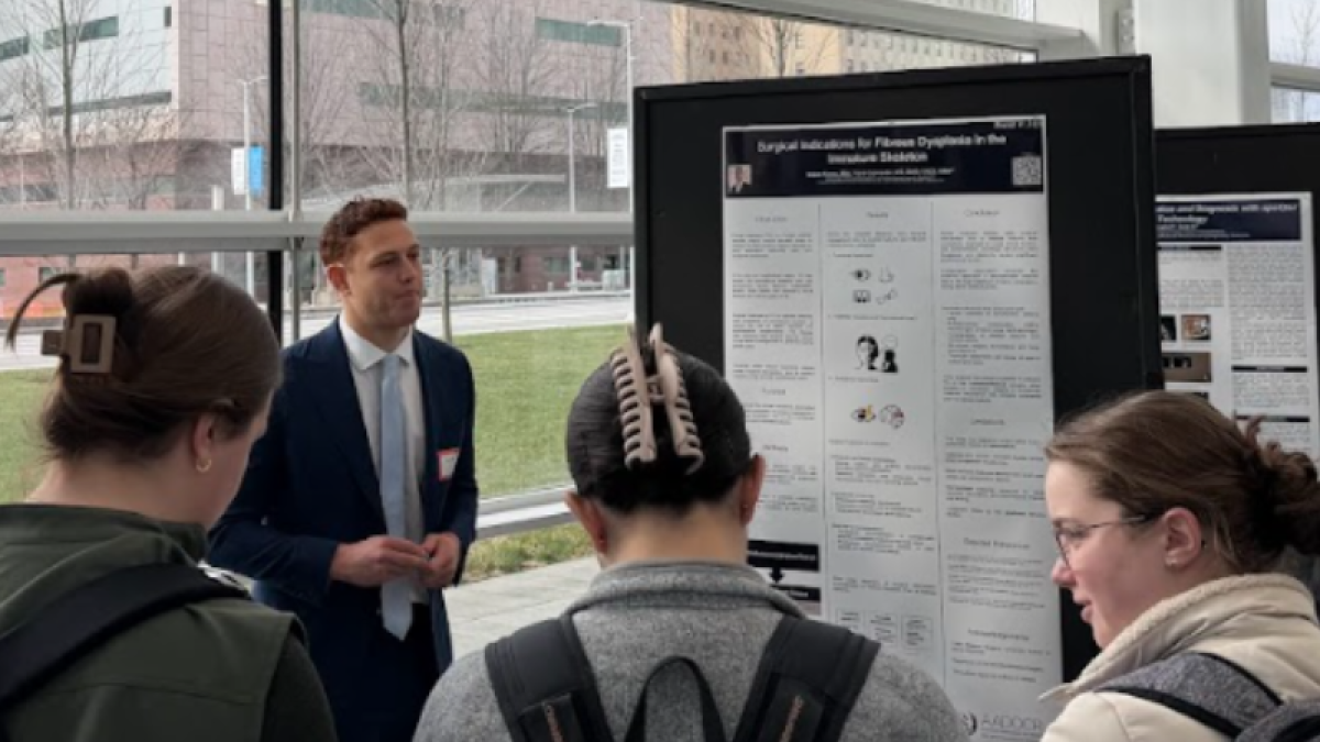 Students gather around a poster presentation being given by a man in a black suit jacket in front of a glass wall.