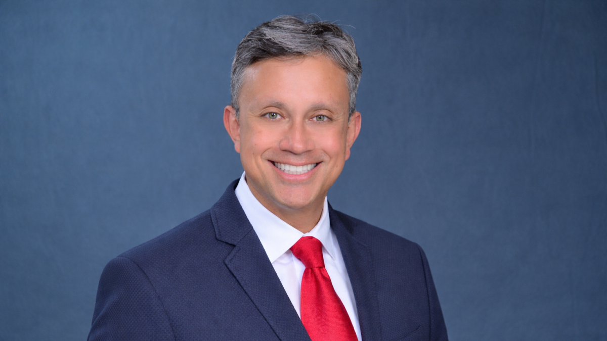 Andres Pinto poses in a blue jacket and red tie in front of a blue background.