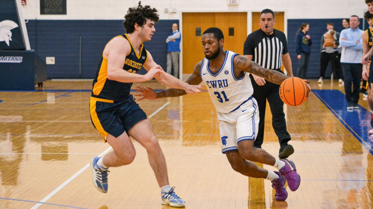 A basketball player in a white CWRU jersey dribbles the ball past a defender in a navy and yellow jersey. A referee observes in the background. 