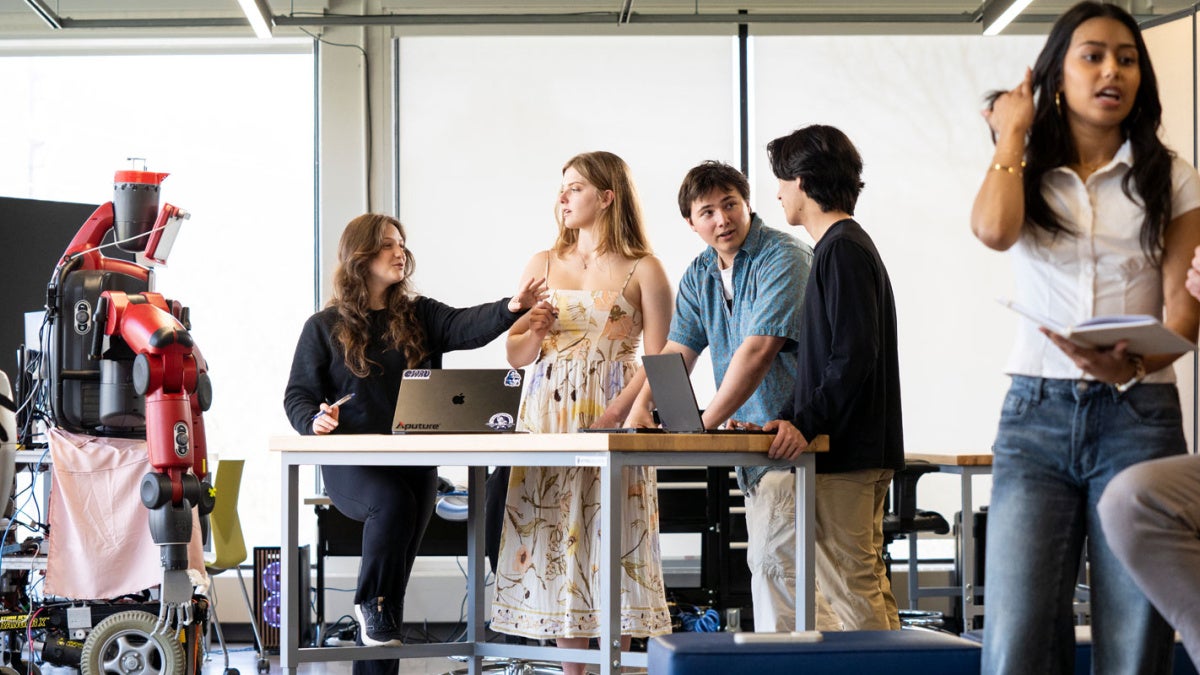 A group of Case Western Reserve University students stand in a classroom, some around a table; robotics are in the corner