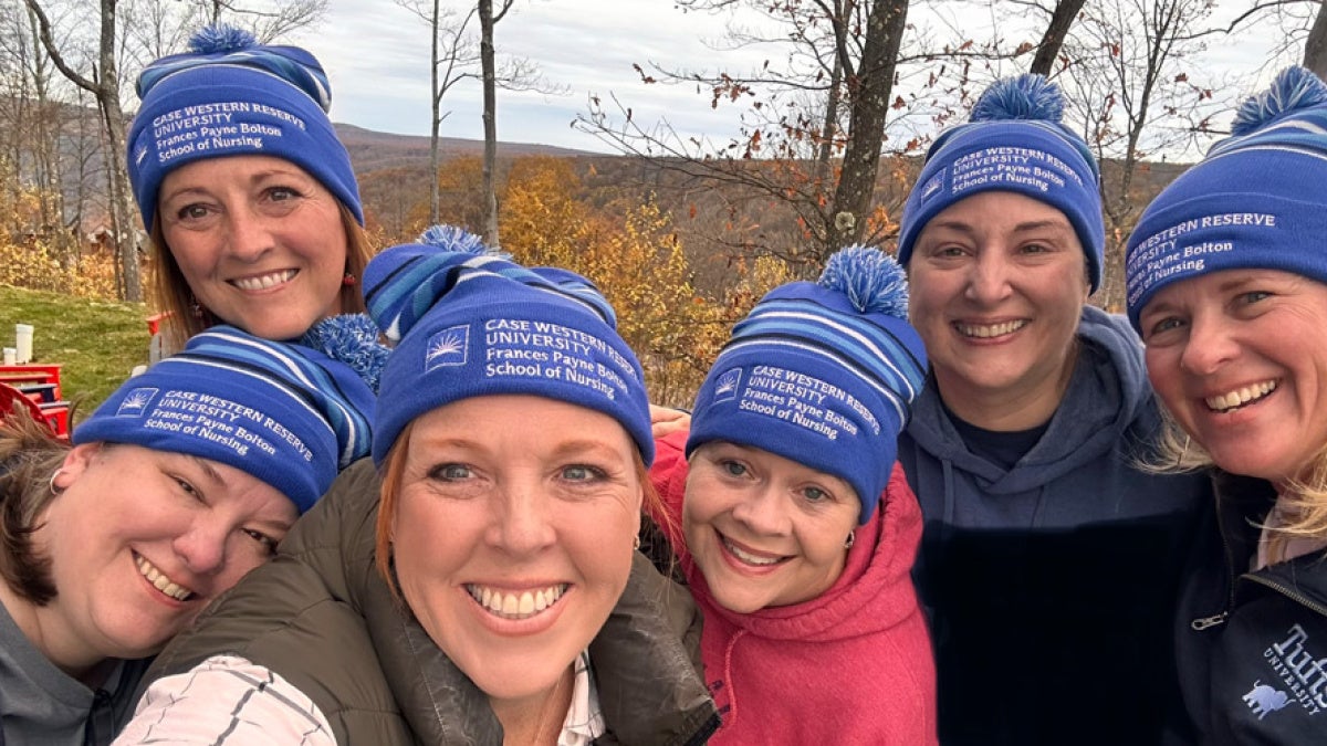 Group of six smiling people in blue beanies from Case Western Reserve University School of Nursing. They're outdoors with a scenic, wooded background.