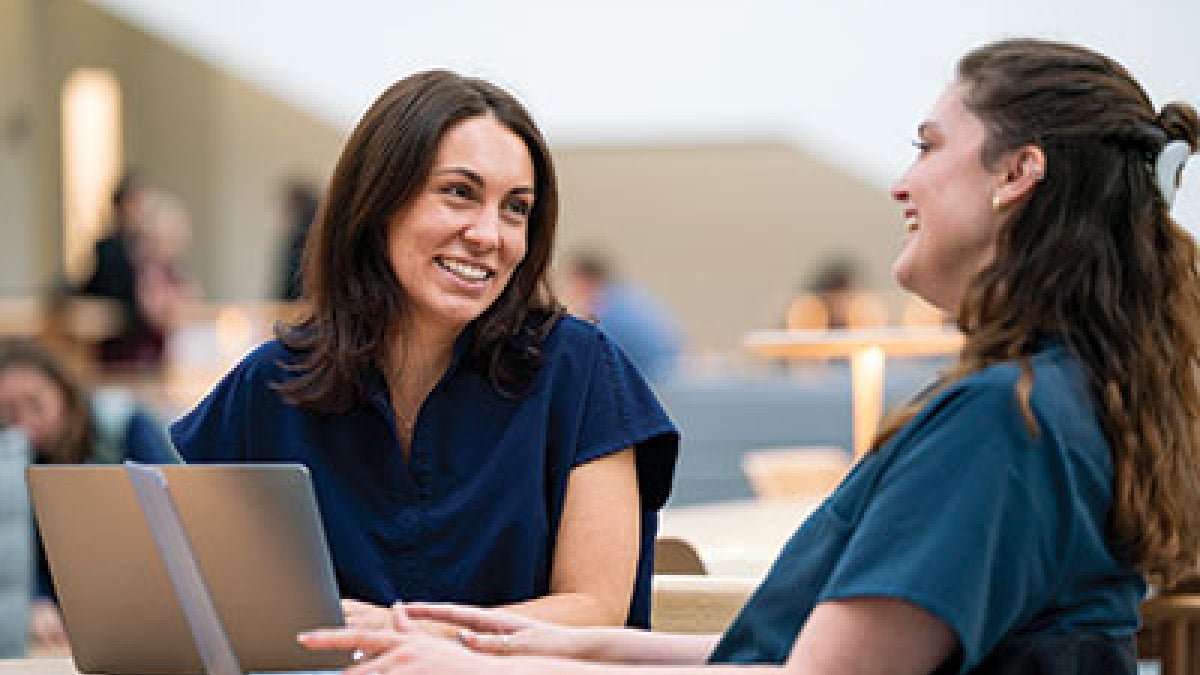 Two women in blue scrubs sit at a table, smiling and talking, with a laptop open in front of them. The setting appears to be a bright, modern space.