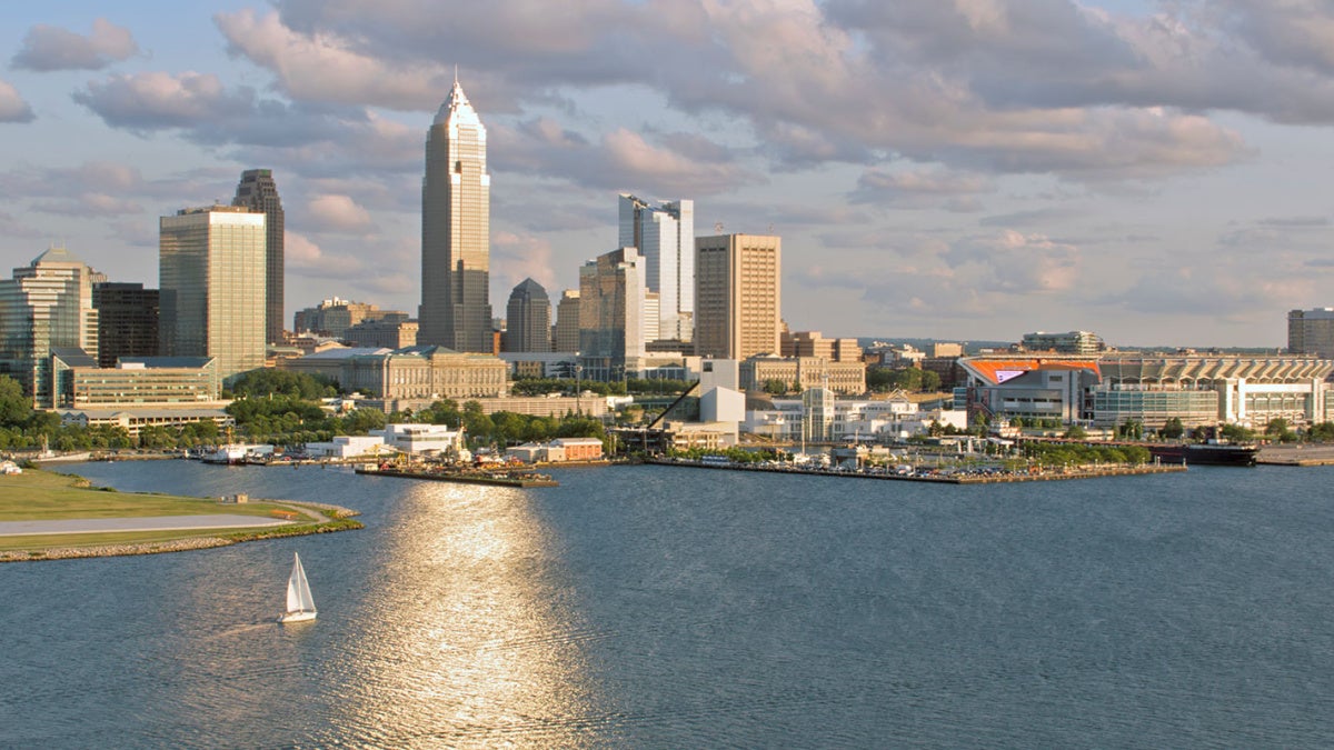 Photo of the Cleveland skyline on a summer day, with the sun reflecting on Lake Erie and a sailboat on the water