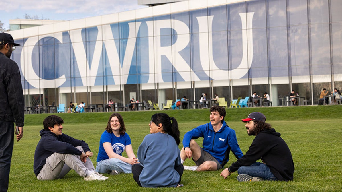 Five students sit in a circle on the ground outside of Tinkham Veale University Center, with large CWRU letters on the windows of the building.