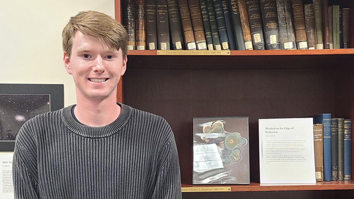 Aidan Selkirk poses for a photo with a copy of his artwork created with an aerosol jet printer framed on a shelf next to him