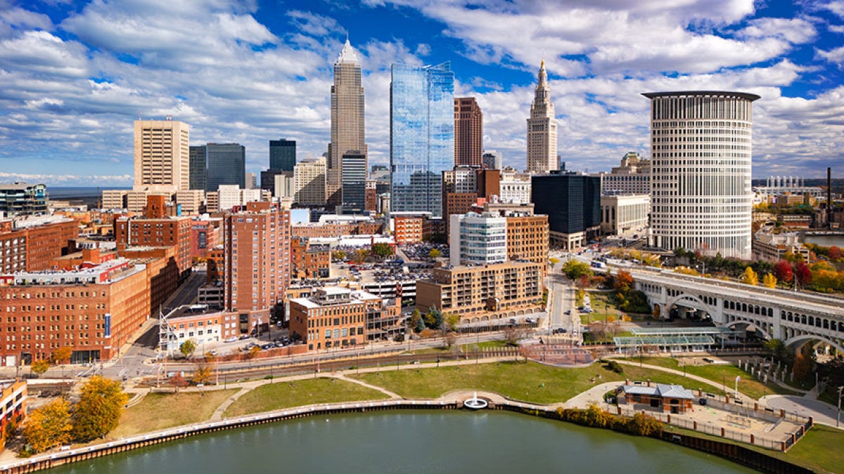 Photo of the Cleveland skyline with clouds set against a bright blue sky and the Cuyahoga River in the foreground