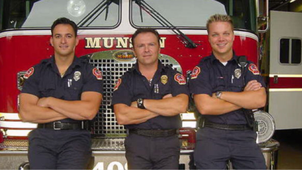 Three men stand in front of a fire truck with arms crossed.