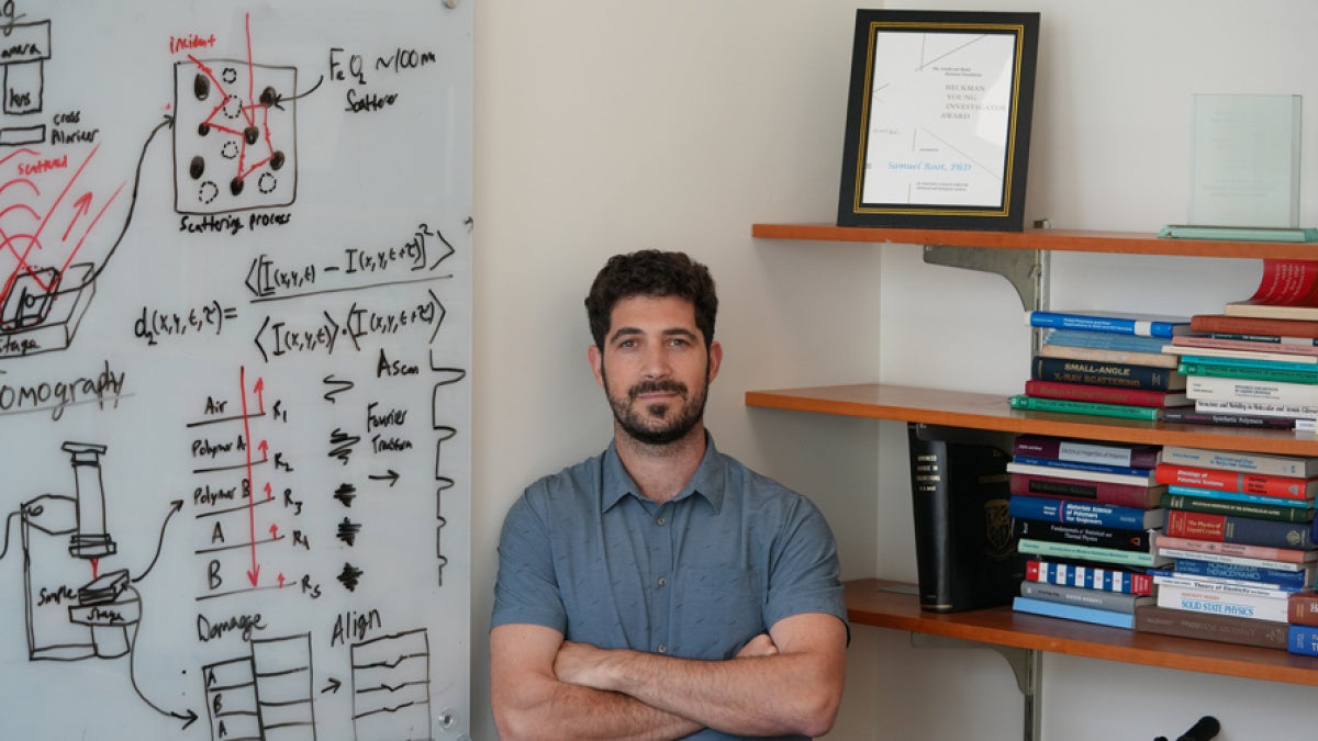 A researcher stands beside a whiteboard covered with diagrams, equations, and notes about imaging, scattering, and polymers. On his right, there are shelves filled with scientific textbooks and framed research awards.