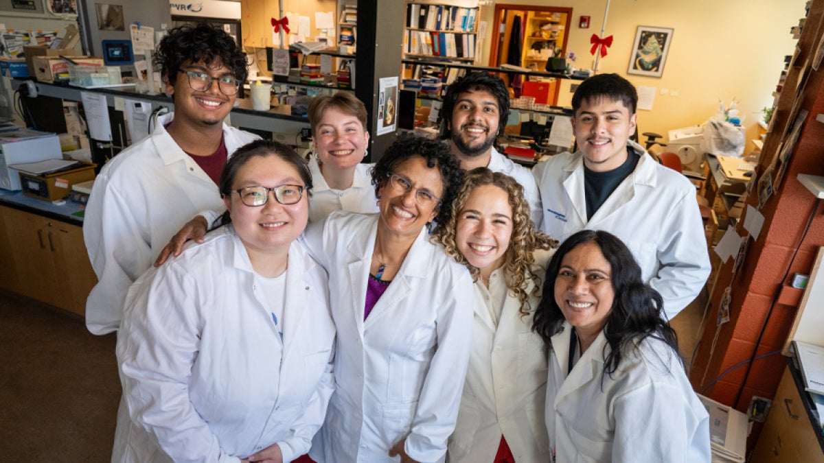 Seven students and a faculty member pose together in white lab coats inside a research laboratory, smiling at the camera. Shelves, equipment, and research materials are visible in the background.