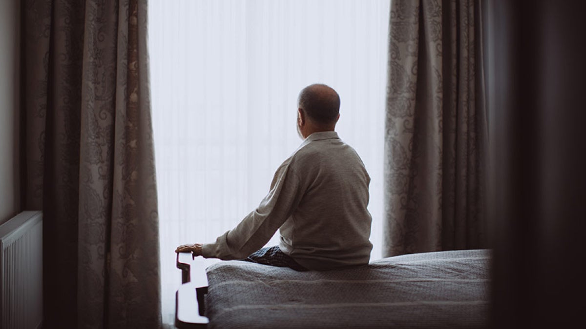 Photo of an elderly man sitting alone on a bed in a darkened room