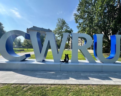 A black, white and brown dog poses in front of a large gray and blue sign near the university’s Binary Walkway that spells, “CWRU.”