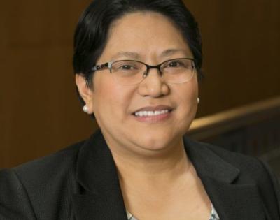 Anna Samia is wearing a black blazer and a patterned blouse, and sitting in front of a wood-paneled background.