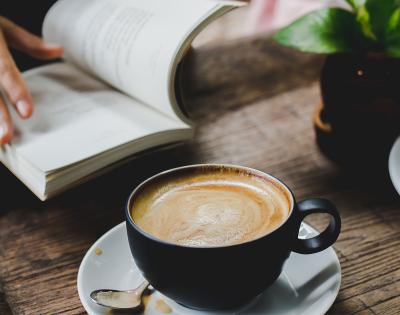 Close-up photo of coffee in a cup on a table and someone reading a book next to it 