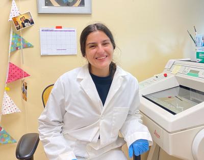 A woman wearing a lab coat and blue gloves smiling in a lab. She is next to scientific equipment.
