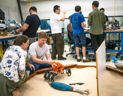 Two people examine a robotic crab on sand, with several other lab workers doing other activities in the background.