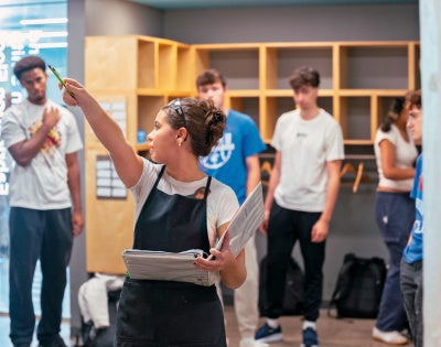 A student in a black apron points with a pen, holding a notebook, in a room with wall cubbies. Several other students watch attentively.