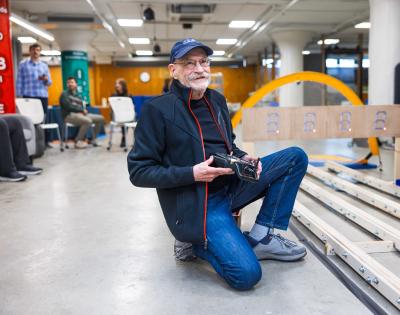 Larry Sears kneels, smiling, holding a remote control with tracks in a space at think[box]. 